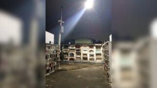 Em primeiro plano, vê-se um corredor pavimentado ladeado por estruturas de túmulos verticais (gavetas funerárias). No centro, um poste alto sustenta um refletor potente aceso, que ilumina intensamente o local, e câmeras de monitoramento instaladas logo abaixo da luminária, apontadas para diferentes direções do pátio. O fundo exibe mais fileiras de túmulos sob um céu noturno escuro.