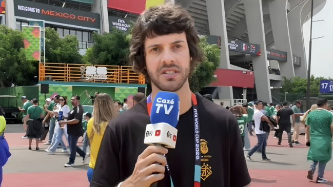 Fotografia colorida em plano médio centralizando o humorista Diogo Defante como repórter de campo. Ele tem pele clara, cabelos castanhos ondulados e barba rala, veste uma camiseta preta com credencial da Copa do Mundo de 2026 no pescoço e segura um microfone azul com a marca da Cazé TV. O cenário ao fundo é a área externa de um grande estádio de futebol durante o dia, onde pessoas circulam vestindo camisas de seleções e há letreiros com as inscrições "Welcome to Mexico City" e "FIFA World Cup 2026".