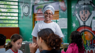 Aqui está a descrição da imagem no estilo "para cego ver", mantendo o padrão breve e direto: Foto em plano médio de um refeitório escolar colorido. No centro, uma merendeira negra sorri para a câmera vestindo camiseta branca, avental e touca descartável na cabeça. Em primeiro plano, três crianças aparecem de costas ou de perfil, focadas nas refeições. O ambiente ao fundo possui paredes verdes decoradas com grafites e desenhos infantis de cozinheiro, garfo e faca. À esquerda, uma janela com grades revela plantas na área externa. A iluminação é difusa e o clima é acolhedor.