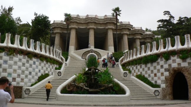 Escadaria principal do Parque Güell em Barcelona, cercada por muros sinuosos em mosaico de cerâmica e colunas neoclássicas ao fundo sob céu nublado.