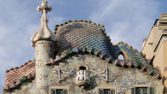 Detalhe superior do telhado sinuoso e colorido da Casa Batlló em Barcelona, imitando as escamas de um dragão com mosaicos e uma torre com cruz.
