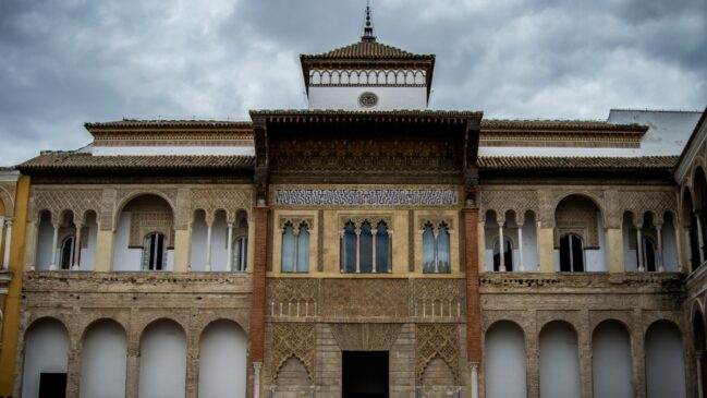 Fachada detalhada em estilo mudéjar do palácio do Real Alcázar de Sevilha, com arcos de ferradura ornamentados sob um céu com nuvens escuras.
