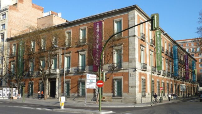 Prédio de tijolos vermelhos e molduras de pedra do Museu Nacional Thyssen-Bornemisza em uma esquina de Madri, com faixas coloridas nas janelas.