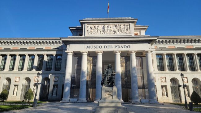 Fachada neoclássica do Museu do Prado em Madri com a estátua do pintor Velázquez em destaque na frente sob céu azul.