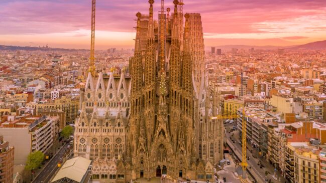 Vista aérea aproximada da Basílica da Sagrada Família em Barcelona, com detalhes de suas torres detalhadas sob um céu de fim de tarde rosa e alaranjado.