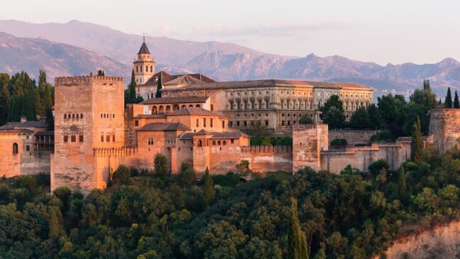 Complexo fortificado do palácio de Alhambra em Granada, situado sobre uma colina verdejante com montanhas ao fundo durante o entardecer