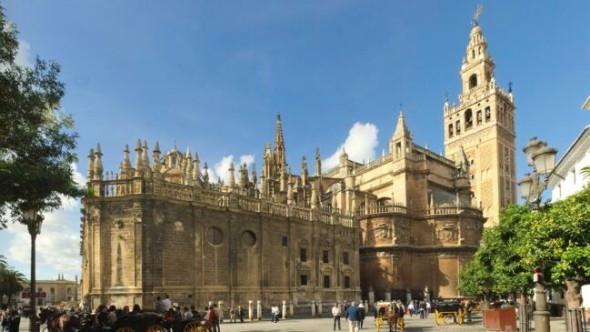 Fachada lateral em pedra da Catedral de Sevilha com a famosa torre de Giralda erguendo-se ao fundo à direita sob céu aberto