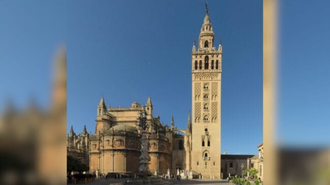 A imponente torre sineira da Giralda erguendo-se ao lado da estrutura gótica da Catedral de Sevilha sob um céu azul limpo.