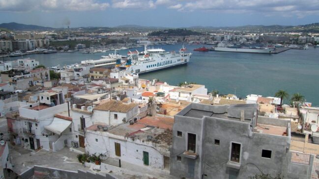 Vista de cima de casas de fachadas brancas na costa de Ibiza com um grande navio de balsa atracado no porto ao fundo