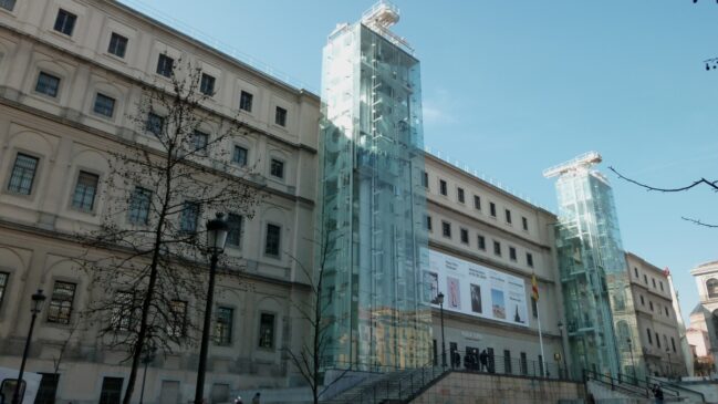 Fachada externa do Museu Nacional Centro de Arte Reina Sofía em Madri, destacando as imponentes torres de elevadores de vidro modernas anexadas ao edifício histórico.