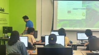 A imagem mostra uma sala de aula ou laboratório de informática com alunos e um instrutor. Em primeiro plano, três alunos aparecem de costas sentados em bancadas de madeira clara diante de monitores de computador: à esquerda, uma pessoa com jaqueta jeans; ao centro, uma pessoa vestindo boné preto com um detalhe amarelo; e à direita, outra de camiseta amarela. Ao fundo, mais à direita, um jovem observa a tela do computador, enquanto à esquerda, um homem de camiseta azul, óculos e cabelo crespo — possivelmente o professor — caminha ao lado das mesas. A parede esquerda do ambiente é pintada de verde-limão com letras pretas, e na parede ao fundo há um grande telão de projeção exibindo janelas de softwares e sistemas operacionais abertas.
