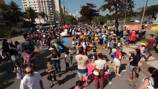 A imagem mostra uma avenida litorânea interditada para carros e completamente ocupada por uma grande quantidade de pessoas em um dia ensolarado. Capturada de um ângulo elevado, a cena revela famílias, adultos e crianças caminhando, conversando e brincando ao longo do asfalto. À esquerda, erguem-se prédios residenciais altos e árvores, enquanto à direita nota-se o início do calçadão com ciclovia, árvores frondosas, alguns guarda-sóis coloridos e um carrinho de sorvete. Há tapetes e lonas coloridas estendidas no chão no centro da pista, onde algumas crianças se concentram para atividades de lazer. O clima geral é de um evento comunitário e recreativo ao ar livre.