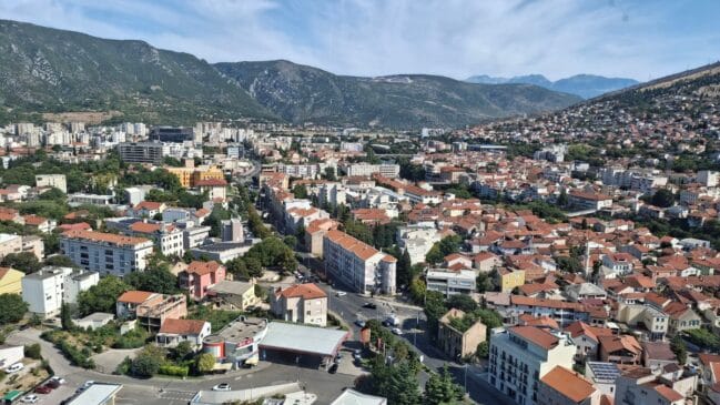 Texto alternativo Fotografia aérea de plano aberto exibindo uma visão panorâmica da cidade de Mostar com seus prédios de telhado avermelhado cercada por montanhas na Bósnia
