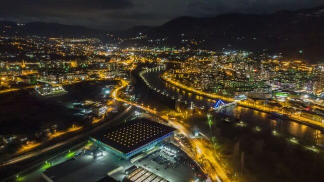 Fotografia aérea noturna exibindo a cidade de Zenica iluminada com o rio Bosna cortando o centro urbano entre montanhas