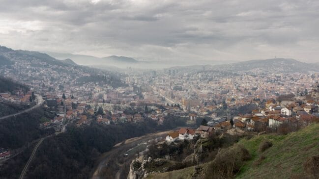 Fotografia horizontal de plano aberto exibindo a vista panorâmica de Sarajevo distribuída pelo vale sob céu nublado