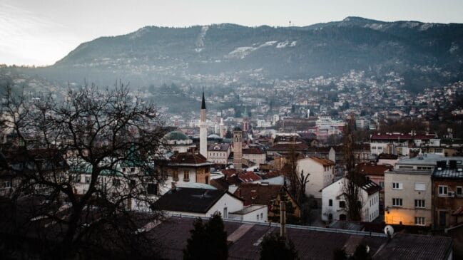 Fotografia horizontal mostrando o panorama dos telhados de Sarajevo com minaretes de mesquitas em destaque diante da montanha