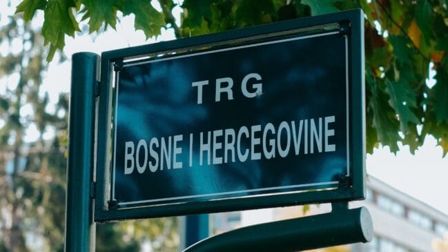 Fotografia em plano detalhe focando uma placa de rua retangular preta com a inscrição em letras brancas Trg Bosne i Hercegovine