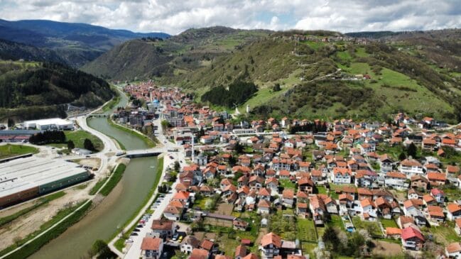 Fotografia aérea de plano aberto exibindo um vilarejo bósnio cortado por um rio sinuoso ao lado de colinas verdes