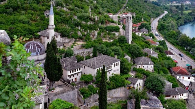 Fotografia horizontal registrando a antiga vila de Počitelj com suas construções de pedra, minarete e torre de vigia entre a vegetação