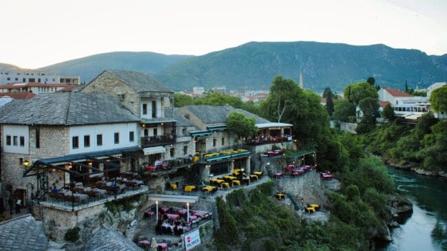 Fotografia horizontal registrando restaurantes e construções de pedra antiga dispostos em patamares na encosta do rio em Mostar