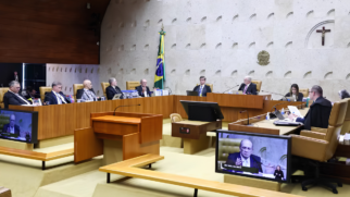 Foto do plenário do Supremo Tribunal Federal durante uma sessão de julgamento. Os ministros aparecem sentados em suas respectivas bancadas de madeira dispostas em semicírculo, vestindo camisas sociais, gravatas e togas pretas. Ao fundo, a parede de mármore claro exibe o Brasão da República, um crucifixo de madeira e a bandeira do Brasil hasteada verticalmente. No plano frontal, mesas de madeira clara organizam o espaço, acompanhadas por dois monitores de vídeo que transmitem o plano fechado de um dos ministros discursando, com o auxílio de uma intérprete de Libras no canto da tela.
