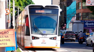 A imagem exibe o VLT prefixo A-003 trafegando pela linha L2 com destino ao Valongo, inserido no fluxo urbano ao lado de carros e de uma motocicleta. A composição visual adota um enquadramento ao nível dos olhos que destaca a identidade visual da Artesp na dianteira branca e laranja do veículo, enquanto placas de sinalização viária e fachadas comerciais ao fundo contextualizam a operação do transporte público na avenida.