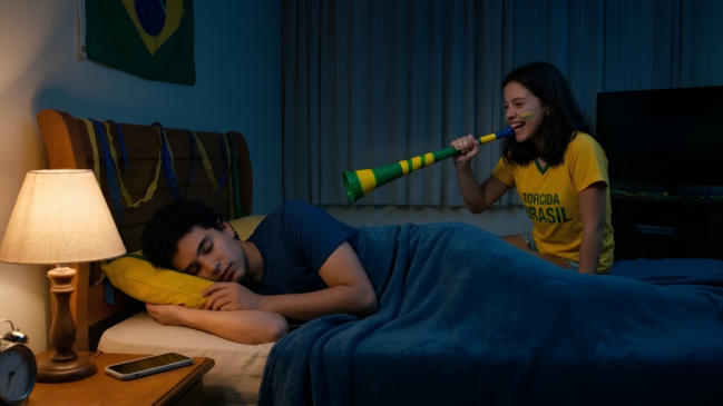 Uma fotografia em plano médio, ambientada em um quarto iluminado suavemente à noite. À esquerda, um homem jovem de cabelos escuros e cacheados dorme deitado de lado em uma cama de casal com cabeceira de madeira. Ele está coberto até a cintura por um edredom azul-escuro, veste uma camiseta azul e apoia a cabeça em um travesseiro amarelo. Na mesa de cabeceira em primeiro plano, há um abajur aceso com cúpula bege, um despertador analógico e um celular deitado. À direita, sentada na cama atrás do homem, uma jovem sorri amplamente enquanto segura uma vuvuzela listrada em verde e amarelo, apontando a abertura em direção ao ouvido do homem. Ela usa uma camiseta amarela com a inscrição verde "TORCIDA BRASIL" e tem detalhes em verde e amarelo pintados na bochecha. O quarto está decorado no clima da Copa do Mundo, com uma bandeira do Brasil na parede e fitas decorativas amarradas na cabeceira da cama. Ao fundo, uma janela com cortinas claras está fechada e uma televisão desligada aparece discretamente à direita.