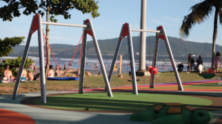 Fotografia de plano médio e ângulo normal de um parquinho infantil à beira-mar, em um dia ensolarado. No primeiro plano, uma escultura colorida em forma de tartaruga, com casco verde e tons de marrom, repousa sobre um piso emborrachado de tons verde-claro e escuro, simulando um jardim. Atrás dela, o chão é colorido com listras amarelas, laranjas e rosas. Ao centro, dominando a composição, duas estruturas metálicas cinzas de balanços se erguem, unidas por suportes superiores laranjas. À esquerda, um balanço com assento circular azul, tipo rede, pende de cordas vermelhas. No suporte à direita, dois balanços menores: um assento vermelho de plástico para bebês e um assento preto simples, pendurados por correntes metálicas. Ao fundo, uma praia movimentada com banhistas e guarda-sóis se estende até o mar calmo. No horizonte, uma grande montanha coberta por vegetação verde e o céu azul-claro com poucas nuvens. Árvores e coqueiros emolduram a cena nos cantos superiores, com pessoas relaxando na grama ao fundo, à direita.