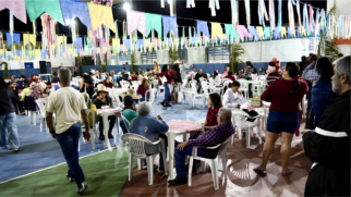A imagem mostra a celebração de uma festa junina noturna realizada em uma quadra poliesportiva coberta. O teto do espaço está todo decorado com fileiras de bandeirinhas juninas coloridas em tons de azul, amarelo, verde e rosa, além de fitas penduradas. No chão da quadra, várias pessoas de diferentes idades estão distribuídas por mesas e cadeiras de plástico branco. Muitas delas vestem trajes típicos, como camisas xadrez e chapéus de palha. Algumas pessoas conversam sentadas ao redor das mesas, que estão cobertas com toalhas xadrez em vermelho e branco, enquanto outras circulam a pé pelo local. Em primeiro plano, à esquerda, um homem de costas caminha usando camiseta branca e calça jeans. Ao centro, um casal conversa sentado em uma mesa: a mulher veste uma blusa vermelha e o homem, uma camisa xadrez roxa. À direita, uma mulher de costas usa blusa vinho e shorts jeans, segurando uma criança no colo que veste uma touca listrada. O ambiente é amplo, bem iluminado e apresenta um clima familiar de confraternização comunitária.