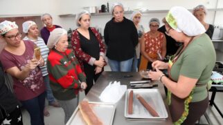 Uma foto horizontal, capturada em ângulo médio, revela um grupo de nove mulheres adultas e idosas, com toucas de cabelo descartáveis, reunidas em uma cozinha de ensino com paredes brancas e piso cinza claro. A maioria das mulheres foca sua atenção em uma mulher mais velha, vestindo uma camiseta verde oliva e avental marrom, que está em primeiro plano, no canto inferior direito, de frente para a direita, demonstrando uma técnica de preparo de chocolate. No centro da imagem, uma bancada de metal segura duas assadeiras de metal. Uma contém uma barra grossa de massa marrom claro polvilhada com cacau em pó, enquanto a outra, à direita, contém um rolo de massa marrom escuro mais estreito, coberto por uma folha transparente. Mais à direita, um pequeno pote de plástico transparente contém um pouco de calda de chocolate marrom. As mulheres que observam têm uma variedade de expressões corporais e faciais. Em primeiro plano, uma mulher com uma camiseta de manga curta roxa escuro com padrão de flores segura um celular dourado na horizontal, possivelmente gravando a demonstração. Uma mulher idosa, à direita dela, com um suéter vermelho, verde escuro e dourado com padrão geométrico, observa atentamente, com as mãos cruzadas na frente do corpo. As outras mulheres, em segundo plano, observam de diferentes posições, uma delas com as mãos na cintura. A cozinha parece bem equipada com utensílios e equipamentos profissionais visíveis nas prateleiras ao fundo. A iluminação é difusa, proveniente de uma fonte de luz superior.