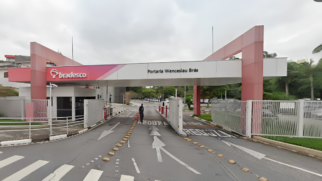 A imagem mostra a entrada de um complexo empresarial ou administrativo do banco Bradesco durante o dia, sob um céu nublado. A estrutura principal da portaria é composta por um grande pórtico horizontal elevado, sustentado por pilares robustos pintados em tom de rosa e vermelho desbotado nas extremidades. A parte central desse pórtico é branca e traz a inscrição em letras pretas "Portaria Wenceslau Brás", enquanto a extremidade esquerda exibe o logotipo redondo e o nome do banco "bradesco" em letras brancas sobre um fundo vermelho. Abaixo da estrutura, há uma via asfaltada de acesso para veículos dividida por tachões amarelos (tartarugas). No asfalto, há setas brancas de sinalização e a palavra "REDUZA" pintada no chão. Um segurança vestindo uniforme escuro caminha de costas pela pista, próximo às cancelas automáticas e portões metálicos brancos que controlam o fluxo de entrada e saída. Do lado esquerdo, há uma pequena guarita de vidro escuro protegida por um corrimão metálico branco e, ao fundo, é possível notar áreas verdes com árvores e um estacionamento com carros distantes.