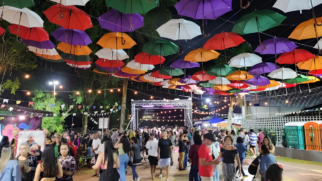 Fotografia colorida e horizontal tirada à noite em um evento ao ar livre. O grande destaque visual está na parte superior, onde dezenas de guarda-chuvas abertos e coloridos (nas cores vermelho, laranja, roxo, verde, branco e amarelo) flutuam suspensos por cabos, cobrindo todo o teto do espaço. Varais com lâmpadas quentes de LED e pequenas bandeirinhas de festa junina cruzam o local sob os guarda-chuvas. Abaixo da decoração, dezenas de pessoas de idades variadas caminham e conversam, preenchendo o calçadão central. Ao fundo, há uma estrutura metálica de palco iluminada e, nas laterais, árvores, barracas e banheiros químicos completam o ambiente festivo.