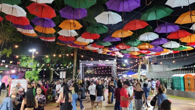 Fotografia colorida e horizontal tirada à noite em um evento ao ar livre. O grande destaque visual está na parte superior, onde dezenas de guarda-chuvas abertos e coloridos (nas cores vermelho, laranja, roxo, verde, branco e amarelo) flutuam suspensos por cabos, cobrindo todo o teto do espaço. Varais com lâmpadas quentes de LED e pequenas bandeirinhas de festa junina cruzam o local sob os guarda-chuvas. Abaixo da decoração, dezenas de pessoas de idades variadas caminham e conversam, preenchendo o calçadão central. Ao fundo, há uma estrutura metálica de palco iluminada e, nas laterais, árvores, barracas e banheiros químicos completam o ambiente festivo.