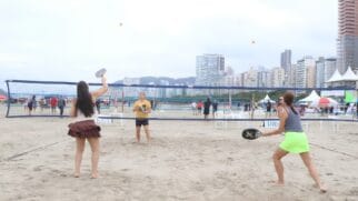 Uma foto horizontal em ângulo médio registra uma partida de beach tennis na areia da praia, sob um céu nublado. Quatro mulheres dividem-se em uma quadra demarcada por linhas pretas na areia, separadas por uma rede azul estendida de ponta a ponta. Em primeiro plano, duas jogadoras estão de costas para a câmera: à esquerda, uma mulher de blusa regata branca e saia azul escura segura a raquete baixa; à direita, outra jogadora veste regata cinza e saia verde-limão, flagrada em movimento com a raquete preta estendida após rebater a bola. Do outro lado da rede, em segundo plano, as outras duas jogadoras posicionam-se de frente: uma à esquerda, de camiseta amarela e bermuda preta, aguarda em postura de defesa; a outra, à direita, veste regata branca e saia marrom, erguendo a raquete preta para rebater uma pequena bola amarela e laranja que flutua no ar perto da rede. Ao fundo, a orla da praia exibe uma fileira densa de prédios altos e uma cadeia de morros sob a névoa. No lado direito, veem-se tendas brancas de eventos armadas na areia, e várias pessoas circulam ou observam o jogo ao redor da rede. A iluminação do dia é clara e difusa.
