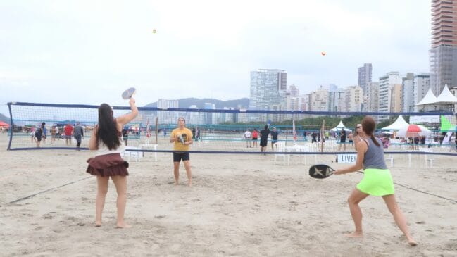 Uma foto horizontal em ângulo médio registra uma partida de beach tennis na areia da praia, sob um céu nublado. Quatro mulheres dividem-se em uma quadra demarcada por linhas pretas na areia, separadas por uma rede azul estendida de ponta a ponta. Em primeiro plano, duas jogadoras estão de costas para a câmera: à esquerda, uma mulher de blusa regata branca e saia azul escura segura a raquete baixa; à direita, outra jogadora veste regata cinza e saia verde-limão, flagrada em movimento com a raquete preta estendida após rebater a bola. Do outro lado da rede, em segundo plano, as outras duas jogadoras posicionam-se de frente: uma à esquerda, de camiseta amarela e bermuda preta, aguarda em postura de defesa; a outra, à direita, veste regata branca e saia marrom, erguendo a raquete preta para rebater uma pequena bola amarela e laranja que flutua no ar perto da rede. Ao fundo, a orla da praia exibe uma fileira densa de prédios altos e uma cadeia de morros sob a névoa. No lado direito, veem-se tendas brancas de eventos armadas na areia, e várias pessoas circulam ou observam o jogo ao redor da rede. A iluminação do dia é clara e difusa.