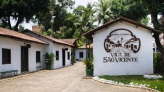 A imagem mostra a entrada e a área externa do Parque Cultural Vila de São Vicente, cenário onde acontecerá o evento mencionado no texto. Em destaque, à direita, há uma parede branca com telhado de barro colonial, exibindo o logotipo oficial do espaço pintado em tons escuros: um brasão estilizado com a inscrição "Parque Cultural Vila de São Vicente". À esquerda, uma alameda pavimentada é margeada por casinhas cenográficas de arquitetura colonial (paredes brancas, portas e janelas de madeira escura), com vasos de plantas decorativos e um banco de praça. Ao fundo e ao redor do complexo, há bastante vegetação, árvores altas e palmeiras sob um céu claro de dia.