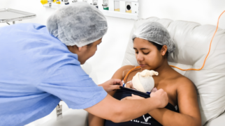 Fotografia colorida e horizontal em ambiente hospitalar de paredes brancas. À direita, uma jovem mãe de cabelos escuros está sentada, reclinada em uma poltrona estofada de cor clara. Ela usa uma touca cirúrgica descartável branca, tem os olhos semicerrados voltados para baixo e segura contra o peito o seu bebê recém-nascido, que veste uma touca de lã clara. A mãe está envolta em uma faixa de tecido escuro onde se lê a inscrição parcial "de São Vicente". À esquerda, em primeiro plano e de perfil, uma profissional de saúde usando uniforme azul e touca descartável branca se inclina em direção à mãe, ajustando com as mãos o tecido que acomoda o bebê. Ao fundo, fixada na parede acima deles, há uma régua hospitalar com saídas de gases medicinais.