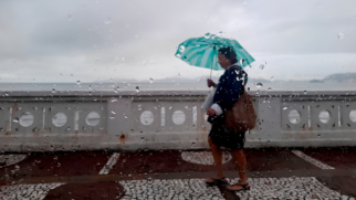 Foto tirada através de um vidro coberto por gotas de chuva, criando um efeito de desfoque na cena externa. No plano de fundo, uma mulher caminha de perfil da direita para a esquerda pela calçada da orla da praia, sob um céu completamente nublado e cinzento. Ela veste casaco escuro, saia, sandálias e carrega uma bolsa marrom no ombro, enquanto segura um guarda-chuva aberto de cor verde-clara. À esquerda, a mureta branca com detalhes vazados vazada divide a calçada do mar, que aparece calmo ao fundo.