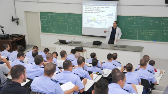 Foto em ângulo plongée de uma sala de aula militar em anfiteatro. Dezenas de alunos fardados com camisas azul-claro anotam a matéria em suas carteiras, vistos de costas. À frente, um instrutor de jaleco branco gesticula em uma bancada de granito. Atrás dele, dois grandes quadros verdes exibem fórmulas manuscritas e, ao centro, uma tela projeta um slide intitulado "SISTEMAS DE DEFESA E VETORES DE ARMAS", ilustrado com o desenho técnico de um caça. O ambiente tem paredes claras e iluminação direta.