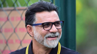 A imagem mostra um plano médio do ex-jogador e comentarista Ricardo Rocha. Ele aparece sorrindo e olhando ligeiramente para a sua direita, fora da direção da câmera. Ele é um homem de pele clara, com cabelos escuros penteados para trás e apresenta uma barba estilosa em tom grisalho, bem aparada ao redor da boca e no queixo. Ele usa óculos de grau com armação retangular escura e veste uma camisa polo preta com listras amarelas finas na gola. O fundo está levemente desfocado, destacando uma estrutura de grade de metal (alambrado) à esquerda e tons de verde e elementos desfocados ao ar livre, sugerindo um ambiente esportivo ou de treinamento.