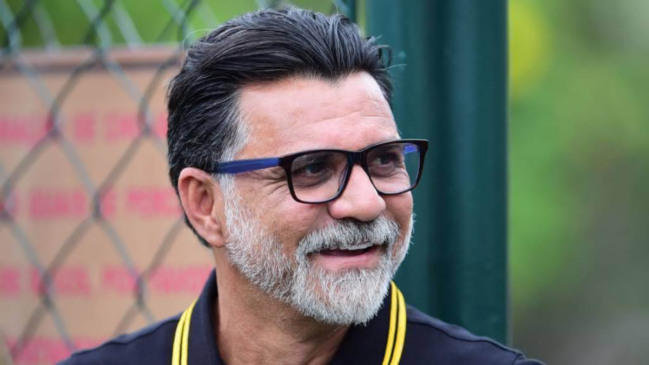 A imagem mostra um plano médio do ex-jogador e comentarista Ricardo Rocha. Ele aparece sorrindo e olhando ligeiramente para a sua direita, fora da direção da câmera. Ele é um homem de pele clara, com cabelos escuros penteados para trás e apresenta uma barba estilosa em tom grisalho, bem aparada ao redor da boca e no queixo. Ele usa óculos de grau com armação retangular escura e veste uma camisa polo preta com listras amarelas finas na gola. O fundo está levemente desfocado, destacando uma estrutura de grade de metal (alambrado) à esquerda e tons de verde e elementos desfocados ao ar livre, sugerindo um ambiente esportivo ou de treinamento.