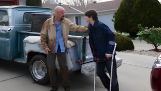 A imagem registra uma cena de forte tensão dramática entre dois personagens da série Breaking Bad, ambientada no interior de uma residência com iluminação quente. Os personagens estão posicionados de perfil, encarando-se diretamente. À esquerda, está o jovem Walter White Jr. (interpretado por RJ Mitte). Ele aparece de perfil para a direita, com uma expressão que mistura surpresa e indignação, marcada pelos lábios entreabertos e o olhar fixo no pai. O jovem tem cabelos castanhos lisos com corte de franja longa, veste uma jaqueta jeans escura com botões metálicos sobre uma camisa xadrez e apoia-se em uma de suas muletas de alumínio com suporte cinza para o antebraço, visível na altura do quadril. À direita, está seu pai, Walter White (interpretado por Bryan Cranston). Posicionado de perfil para a esquerda, ele é um homem mais velho, calvo, que usa óculos de grau com armação fina e exibe sua característica fisionomia séria com cavanhaque bem desenhado. Ele veste uma jaqueta leve de sarja em tom bege ou caramelo e está com as duas mãos erguidas à frente do corpo, com os dedos gesticulando abertos no meio de uma fala argumentativa ou defensiva. O plano de fundo mostra o interior da casa da família White, com paredes em tons de ocre, detalhes em madeira no teto e uma janela coberta por uma cortina branca translúcida que suaviza a entrada de luz externa. No canto esquerdo inferior, parte de um móvel de madeira de uma cristaleira também fica visível.