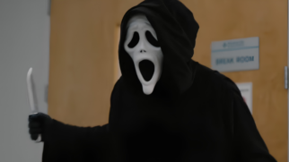 A imagem mostra uma pessoa fantasiada como o icônico vilão Ghostface, personagem da franquia de terror e comédia. O indivíduo está posicionado em plano médio, do centro para o lado direito do enquadramento, voltado levemente para a esquerda. O vilão usa a clássica máscara branca de formato alongado com expressão de pânico, caracterizada pelas cavidades oculares pretas e caídas, dois pequenos pontos pretos no nariz e uma grande boca preta aberta em um grito vertical. A cabeça está coberta pelo capuz de uma túnica inteiramente preta que envolve todo o seu corpo. O braço direito do personagem está flexionado para a frente e para o lado esquerdo da imagem, com a mão vestindo uma luva preta e empunhando uma faca de lâmina branca e cabo escuro, apontada para cima. O plano de fundo exibe um ambiente interno com uma parede branca à esquerda e uma porta de madeira clara no centro e à direita. Fixada na porta, há uma placa retangular cinza com letras azuis onde se lê "BREAK ROOM" (sala de descanso). A iluminação do cenário é difusa e uniforme.