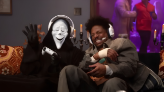 A imagem mostra uma cena cômica e de tom descontraído ambientada em uma sala de estar escura. Duas figuras estão sentadas lado a lado em um sofá preto, jogando videogame. À esquerda, está o assassino mascarado **Ghostface**, vestindo seu clássico manto preto com capuz. Ele usa uma variação da famosa máscara branca — desta vez com uma expressão alegre, olhos fechados em formato de arco e um sorriso largo. O Ghostface usa um headset gamer branco sobre o capuz, segura um controle de videogame branco na mão esquerda e acena para a câmera com a mão direita, que está vestida com uma luva preta. À direita, está um homem negro com cabelo estilo afro curto e barba por fazer, expressando grande entusiasmo ou frustração cômica enquanto joga, com os olhos fortemente cerrados e os dentes à mostra. Ele veste uma jaqueta de sarja cinza sobre uma camiseta de cor clara e tem uma faixa ou atadura branca enrolada no pulso direito. Ele também usa um headset gamer branco com detalhes azuis e segura um controle de videogame preto. O cenário de fundo tem uma iluminação suave e quente proveniente de castiçais com velas acesas posicionados atrás do sofá. No canto superior direito, há uma abertura para outro ambiente com iluminação roxa e neon, onde figuras borradas parecem estar em movimento. Nas extremidades do sofá, almofadas com estampas florais e ornamentos em tons de vermelho e dourado completam o ambiente.