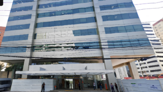 A imagem mostra a fachada de um moderno edifício comercial espelhado, fotografado de um ângulo de baixo para cima durante o dia. O prédio possui uma estrutura de placas cinza-claras intercaladas por amplas fileiras horizontais de janelas de vidro azulado, que refletem levemente o céu. Cruzando horizontalmente toda a extensão da imagem, na metade inferior da fachada, há uma grande quantidade de cabos e fios de energia pretos que passam pela rua. Na base do edifício, há uma entrada recuada com portas de vidro, protegida por uma marquise branca proeminente. Tapumes metálicos brancos cercam as laterais da calçada. Poucas pessoas circulam ou estão paradas em frente à entrada, incluindo o que parece ser um segurança à esquerda e pedestres conversando no lado direito. À extrema esquerda, a lateral de uma van cinza está parcialmente visível.