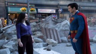 Fotografia colorida em plano médio mostrando um homem fantasiado de Superman e uma mulher conversando em uma rua urbana tomada por escombros. Primeiro plano: No lado esquerdo, uma mulher de pele clara e longos cabelos pretos sorri levemente enquanto olha para a direita. Ela veste um casaco de tricô roxo de botões, calça escura de cintura alta com cinto e um relógio de pulso dourado. No lado direito, um homem de pele clara e cabelos pretos curtos está posicionado de perfil, olhando para ela. Ele usa o traje clássico do Superman, composto por um macacão azul justo com o símbolo do herói no peito, uma capa vermelha longa fixada nos ombros e um cinto amarelo. Segundo plano: Entre os dois personagens, há um grande amontoado de destroços, pedaços de concreto, poeira e vigas de metal retorcidas espalhadas pelo chão. Ao fundo, observa-se a fachada de prédios comerciais com comércios fechados, letreiros antigos e toldos verdes, além de figurantes distantes caminhando pela calçada sob uma iluminação diurna suave.