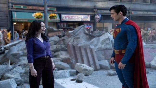 Fotografia colorida em plano médio mostrando um homem fantasiado de Superman e uma mulher conversando em uma rua urbana tomada por escombros. Primeiro plano: No lado esquerdo, uma mulher de pele clara e longos cabelos pretos sorri levemente enquanto olha para a direita. Ela veste um casaco de tricô roxo de botões, calça escura de cintura alta com cinto e um relógio de pulso dourado. No lado direito, um homem de pele clara e cabelos pretos curtos está posicionado de perfil, olhando para ela. Ele usa o traje clássico do Superman, composto por um macacão azul justo com o símbolo do herói no peito, uma capa vermelha longa fixada nos ombros e um cinto amarelo. Segundo plano: Entre os dois personagens, há um grande amontoado de destroços, pedaços de concreto, poeira e vigas de metal retorcidas espalhadas pelo chão. Ao fundo, observa-se a fachada de prédios comerciais com comércios fechados, letreiros antigos e toldos verdes, além de figurantes distantes caminhando pela calçada sob uma iluminação diurna suave.