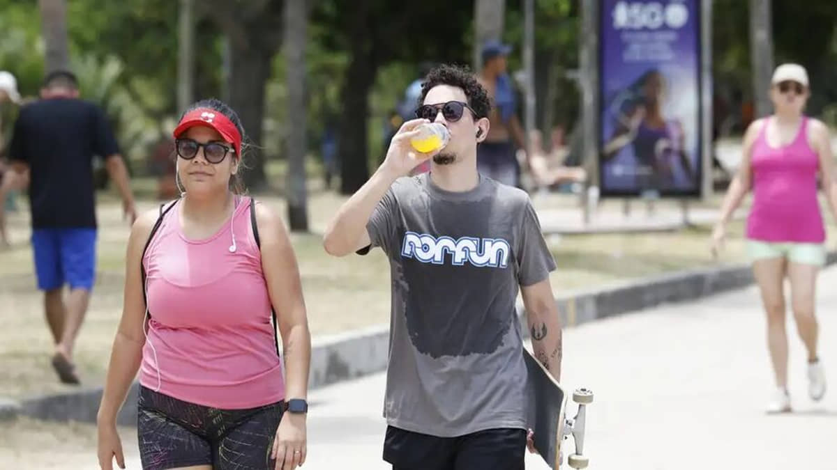 Casal caminha na orla e rapaz bebe água em dia muito quente no litoral