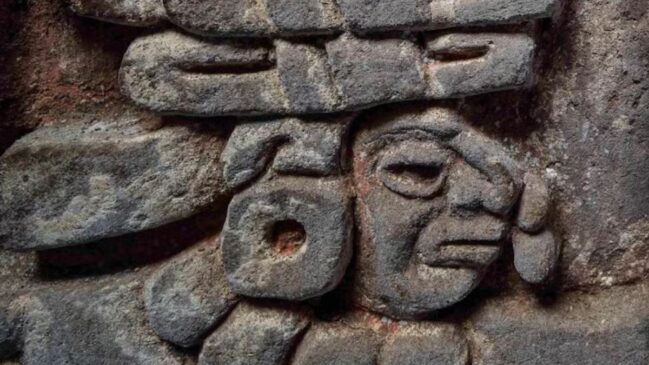 Tesouros do tempo: arqueólogos revelam pinturas murais vibrantes e inscrições do calendário zapoteca em uma tumba datada de 600 d.C. O bico de uma coruja esculpida cobre o rosto de uma figura humana, reforçando o simbolismo espiritual deste local sagrado. / Gerardo Peña/INAH